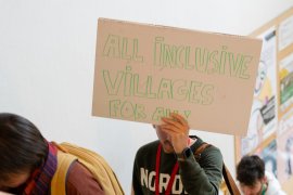 man holding protest sign 'ALL INCLUSIVE VILLAGES FOR ALL!'