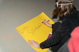 A person writes 'With Solidarity' on a yellow piece of paper while wearing a white eye mask