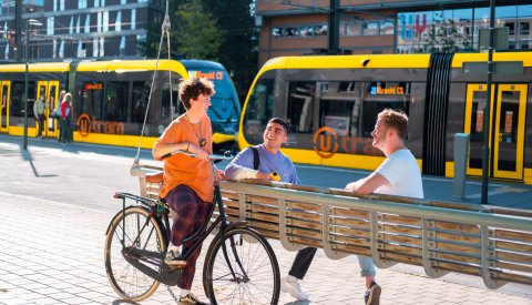 een meisje staat met haar fiets bij een bankje, op het bankje zitten twee jongens die naar haar kijken. achter hun rijdt de tram.