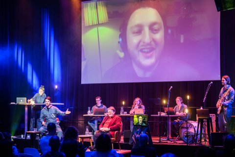 Acht theathermakers, muzikanten en wetenschappers staan op het podium in TivoliVredenburg. Op het grote scherm is nog een persoon te zien.
