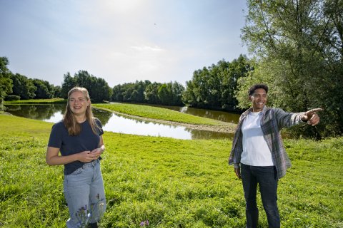 A girl and a boy are standing in a sunny nature reserve. The girl smiles past the camera and has her hands crossed. The boy smiles the other way and points his finger forward.