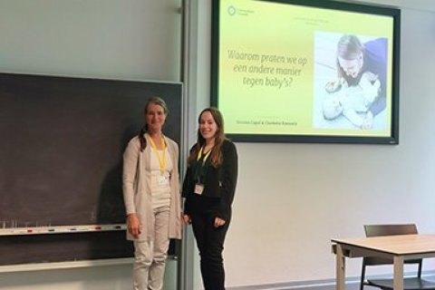 Charlotte Koevoets en Desiree Capel poseren voor het scherm met daarop hun PowerPoint-presentatie. Op het scherm is een moeder met baby te zien en de tekst: 'Waarom praten we op een andere manier tegen baby's?'