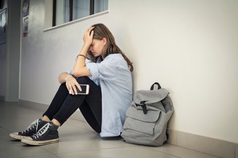 Een tienermeisje zit verdrietig tegen een muur met een schooltas naast zich.