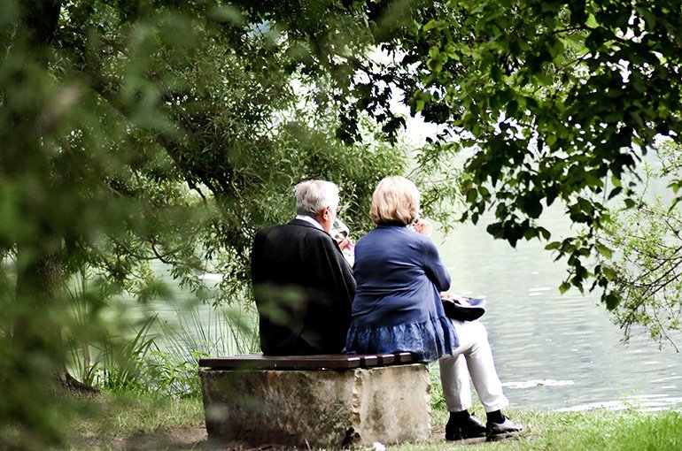 Twee senioren die witte wijn drinken op een bankje in de natuur. Foto: Sven Mieke, via Unsplash