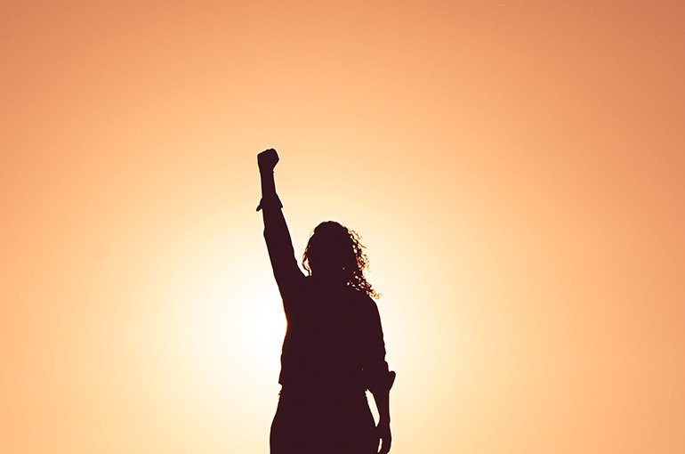 Silhouette van een vrouw met vuist in de lucht. Foto: Miguel Bruna, via Unsplash