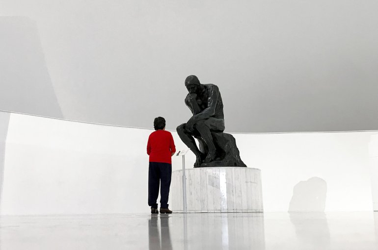 Museum visitor wearing a red sweater stands in front of Rodins The Thinker in a white space